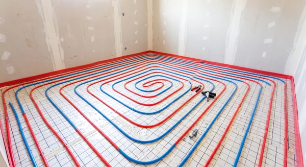 Chantier intérieur en construction, pièce vide avec murs en plaques de plâtre partiellement enduites. Sol recouvert de panneaux isolants quadrillés à plots. Tubes PER (polyéthylène réticulé) de couleur bleu ou rouge disposés en serpentin régulier sur la surface isolante, fixés par agrafes ou clips. Bande de désolidarisation rouge visible en bordure périphérique. Espacement régulier entre les tubes caractéristique d'une installation de plancher chauffant hydraulique neuve. Ambiance de chantier professionnel en cours d'exécution.
