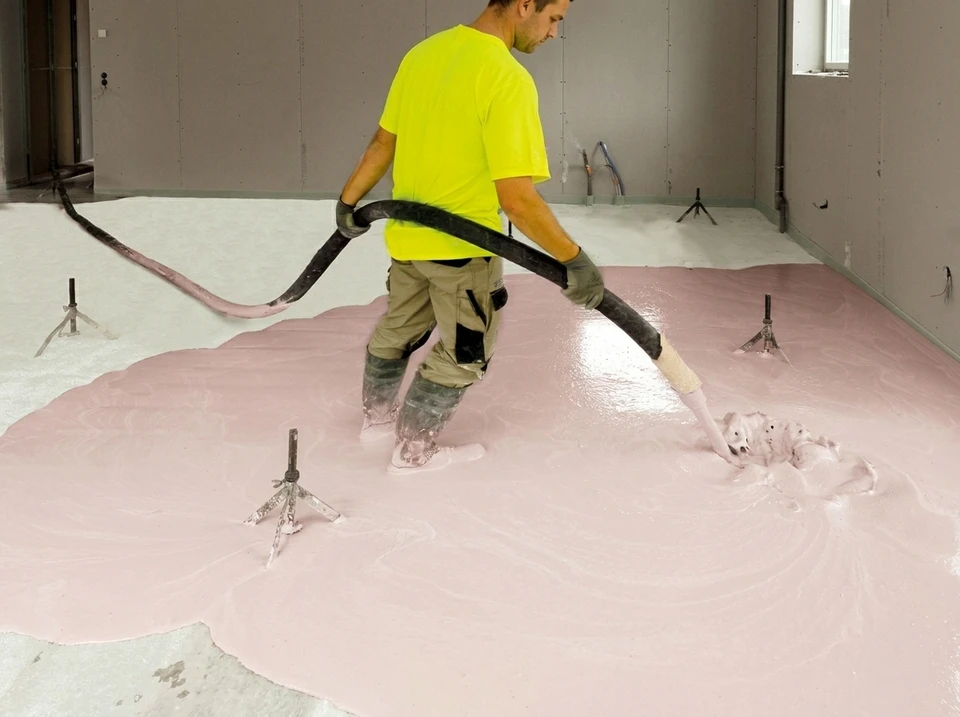 Technicien en gilet jaune fluo appliquant une chape fluide autonivelante rose via un tuyau flexible noir dans un local en construction